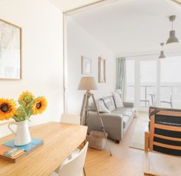 A living room with a wooden dining table and sunflowers in a vase next to a gray sofa and armchair with large windows overlooking the sea at 12 West End Point in Pwllheli