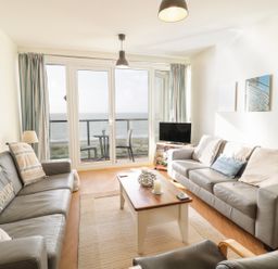 A living room with two sofas, a coffee table, a television on a stand, and a balcony with a table and two chairs overlooking the ocean at 12 West End Point in Pwllheli