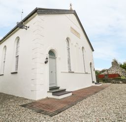 Exterior view of a white building with arched windows and doors surrounded by gravel and a brick pathway with another stone building in the background at Capel Bryn Bachau Chwilog near Criccieth