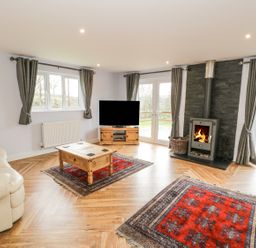 A living room with a cream sofa a wooden coffee table a TV on a wooden stand two red rugs and a wood burning stove at Nant Y Felin Lodge in Aberdaron