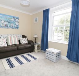 A living room with a brown sofa with cushions and a small table with a lamp beside a window with blue curtains at No 9 in Watchet