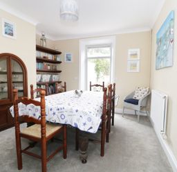 A dining room with a wooden table covered by a patterned tablecloth surrounded by wooden chairs a glass-fronted cabinet shelves with books and decorative items a gray armchair with cushions and framed pictures on the walls at No 9 in Watchet