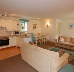 An open plan living room with two beige sofas a green rug a wooden dining table with chairs and a kitchen area with white cabinets at The Coach House in Ugborough
