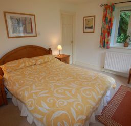 A bedroom with a double bed two bedside tables with lamps framed pictures a window with curtains and a chair at The Coach House in Ugborough