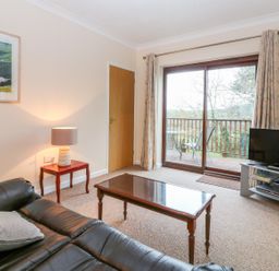 A living room with a leather sofa a wooden coffee table a lamp on a side table a television on a stand and sliding glass doors leading to a balcony at Little Waycroft in Dawlish