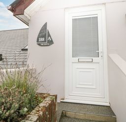 A white door with a sailboat shaped house number plaque next to a brick planter with plants at Monkbarns in Brixham