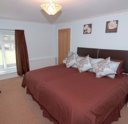 A bedroom with a large bed with brown bedding and multiple pillows bedside tables with lamps a chair and a window with brown curtains at Garden View in Kingskerswell