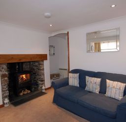 A living room with a blue sofa and three striped cushions a fireplace with burning wood and stone surround and a large mirror on the wall at The Keep Pot in Port Isaac