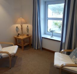 A small room with two white cushioned wooden chairs a wooden side table with a lamp and decorations and a window with curtains at The Keep Pot in Port Isaac
