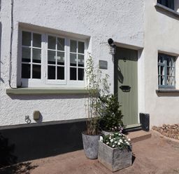 Exterior of a white stucco cottage with green door and two windows with flower pots outside at Robyn Cottage Otterton