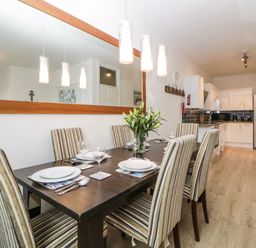 A dining area with a dark wooden table and six striped chairs set with plates and glasses next to a kitchen with white cabinets and appliances at Quayside Lookout in Brixham