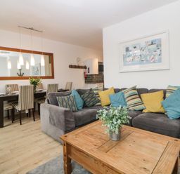 A living room with a grey sectional sofa with colorful cushions a wooden coffee table with a plant and a dining area with a table and striped chairs at Quayside Lookout in Brixham