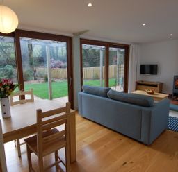 A living room with a wooden dining table and chairs a blue sofa a wood burning stove and large glass doors overlooking a garden at Orchard Retreat in Sidmouth