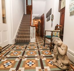 A hallway with patterned tiled floor a striped carpeted staircase a wooden chair with cushions a statue a grandfather clock and wall signs at Muriau Park in Caernarfon