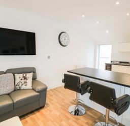 A living area with a gray sofa with animal print cushions a wall mounted TV a clock two black bar stools and a kitchen counter at 7 Marina View in Pwllheli