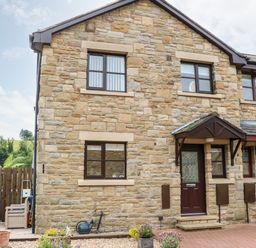 The exterior of a stone house with brown framed windows a dark brown door with a glass panel and a garden area with plants at 14 The Maltings in Rothbury