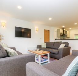 A living room with three gray sofas a wooden coffee table a wall mounted TV and a kitchen in the background at Beech in Truro