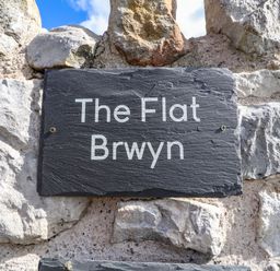 A stone wall with a dark plaque reading The Flat Brwyn at The Flat At Brwyn in Brynford