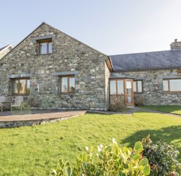 A stone house with wooden framed windows and a stone patio with chairs surrounded by grass at Awel Y Llan in Llanfair
