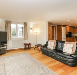 A living room with a black leather sofa a large TV on a stand a rug wooden floor and a view of a kitchen with wooden cabinets at The Annex in Royal Wootton Bassett