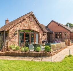 An outdoor patio area with glass fencing and garden bushes by a stone house at The Old Byres in Washford near Watchet