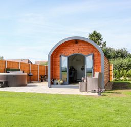 A wooden pod with open doors on a patio with chairs and a hot tub in a fenced garden at Withy Willows in Diddlebury near Ludlow