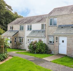 A row of stone and brick terraced houses with white doors and windows along a paved path with green lawns and shrubs Oyster Cottage in Falmouth