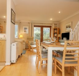 A dining area with a wooden table and chairs set with plates and glasses next to a living room with armchairs and a bookshelf at Oyster Cottage in Falmouth