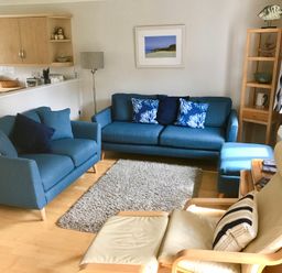A living room with blue sofas a cream armchair a wooden shelving unit and a kitchen pass-through at Oyster Cottage in Falmouth