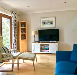 A living room with a blue sofa a beige armchair and ottoman a wooden bookshelf and a TV on a white stand at Oyster Cottage in Falmouth