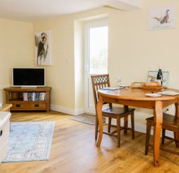 A room with a wooden dining table, two chairs, a small TV on a wooden stand, and animal paintings on the wall at Woodpecker Lodge Henley near Langport