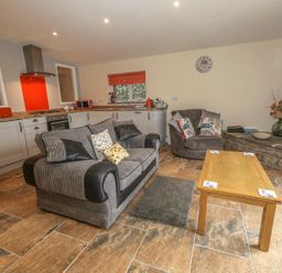A living room with a gray sofa and armchair a wooden coffee table and kitchen area with white cabinets at Knott Lodge in Sedbergh