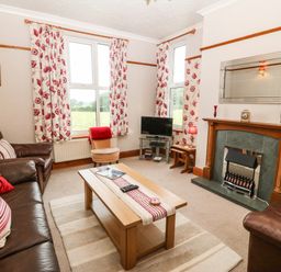 A living room with two windows floral curtains a glass TV stand a wooden coffee table and a fireplace at Dinas Farmhouse Annex in Caernarfon