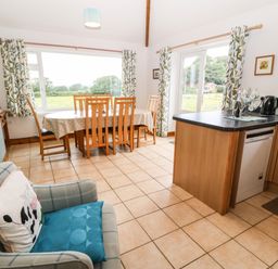 A kitchen dining area with wooden table and chairs next to large windows and a kitchen counter with appliances at Dinas Farmhouse Annex in Caernarfon