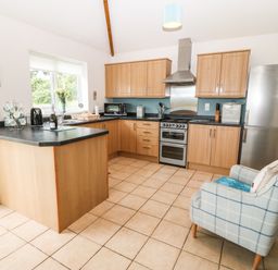 A kitchen with wooden cabinets a stove refrigerator a countertop with a kettle and cups and a checkered armchair near a radiator at Dinas Farmhouse Annex in Caernarfon