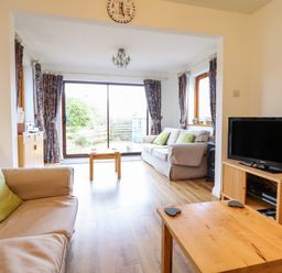 A living room with two beige sofas and green cushions a wooden coffee table a television on a wooden stand and sliding glass doors at Dehangfa in Fairbourne
