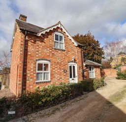 A brick house with white framed windows and a white door on a gravel driveway with bushes around at Elder Cottage in Mickleton near Chipping Campden