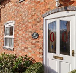 Exterior view of a brick house with a white door featuring stained glass panels and a window with curtains at Elder Cottage in Mickleton near Chipping Campden