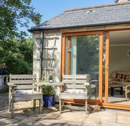 A patio with wooden chairs and a small table with a bottle and glasses outside a house with sliding glass doors showing a living room at Manege Cottage Near St Agnes