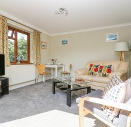 A living room with a beige sofa and patterned cushions a glass coffee table a TV on a stand and a small dining table with two chairs near the window at Manege Cottage Near St Agnes