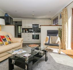 A living room with a beige sofa and patterned cushions a glass coffee table a grey armchair with two cushions and an open door to a garden at Manege Cottage Near St Agnes
