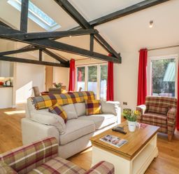 A living room with beige sofa, plaid armchairs, wooden coffee table, red curtains, and exposed black ceiling beams at Zeal Cottage in Crediton