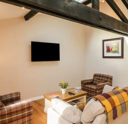 A living room with a wall-mounted TV, two plaid armchairs, a beige sofa with a striped throw, a wooden coffee table with magazines and a vase of flowers, and a framed painting on the wall at Zeal Cottage in Crediton
