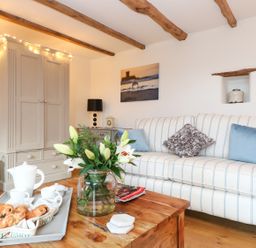 A living room with a striped sofa, wooden beams on the ceiling, a cabinet with string lights, a coffee table with a flower vase and a tea set at Polly's Bower in Landrake