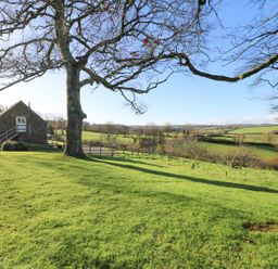 A grassy yard with a large tree and a stone building surrounded by fields and fences at Pollys Bower in Landrake