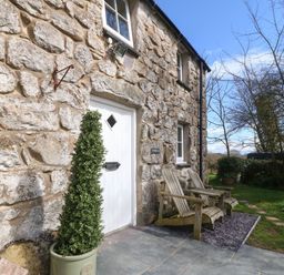 Exterior view of a stone cottage with a white door two wooden chairs and a potted plant at Y Bwthyn Ty Du in Pwllheli