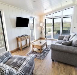 A living room with a grey sofa, plaid armchair, coffee table, TV on the wall, and sliding glass doors at Woodpecker Lodge in Gargrave