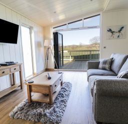 A living room with a grey sofa a wooden coffee table a TV on the wall and a door opening to a deck with a view at Woodpecker Lodge in Gargrave