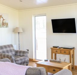 A sitting area with a plaid armchair a floor lamp a wooden TV stand with electronics a wall-mounted flat screen TV and a coffee table at Woodpecker Lodge in Gargrave