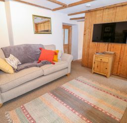 A living room with a sofa with cushions and a throw a patterned rug a wall mounted television and a wooden cabinet at Blue Anchor in Fraddon